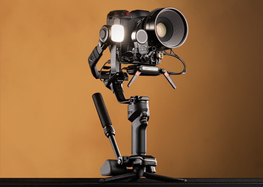 Gimbal ZHIYUN Crane 4 Combo Stabilizator z zamocowanym aparatem ustawiony na statywie. Na urządzeniu znajduje się wiele dodatkowych akcesoriów, spust, pokrętło