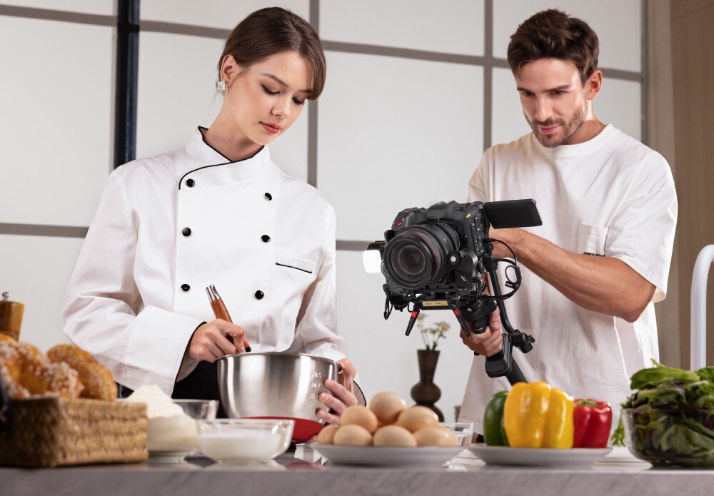 Gimbal ZHIYUN Crane 4 Combo Mężczyzna w białej koszulce trzyma aparat na stabilizatorze, nagrywając kobietę w stroju kucharskim, która miesza składniki w metalowej misce, działanie, cechy, zalety