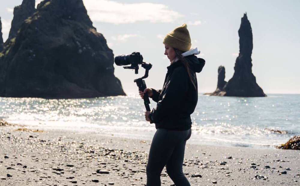 Gimbal ZHIYUN Crane 4 Mężczyzna w czarnym podkoszulku trzyma aparat zamocowany na stabilizatorze, pochylając się lekko do przodu, opis