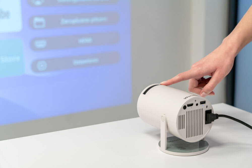 Projektor WANDOO LMP360 HD (1280 x 720), 160 ANSI lumen, Wi-Fi, Bluetooth Projektor Wandoo rzutuje menu na jasną ścianę. Ręka dotyka górnej części obudowy projektora, prawdopodobnie w celu obsługi dotykowego panelu sterującego. Obsługa dotykowego panelu sterowania projektora Wandoo.