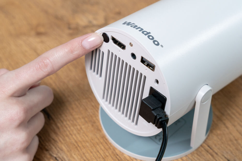 Projektor WANDOO LMP360 HD (1280 x 720), 160 ANSI lumen, Wi-Fi, Bluetooth Ręka wskazuje palcem włącznik zasilania (przycisk) na tylnym panelu projektora Wandoo, obok portów HDMI i USB. Włączanie projektora Wandoo przyciskiem z tyłu obudowy.