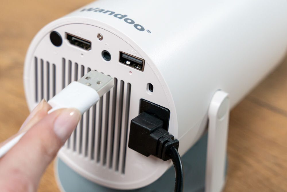 Projektor WANDOO LMP360 HD (1280 x 720), 160 ANSI lumen, Wi-Fi, Bluetooth Zbliżenie na tylny panel białego projektora Wandoo z wlotami powietrza. Ręka podłącza biały kabel USB do portu, obok widoczne są gniazda HDMI, audio i zasilania. Podłączanie pendrive do portu USB w projektorze Wandoo.