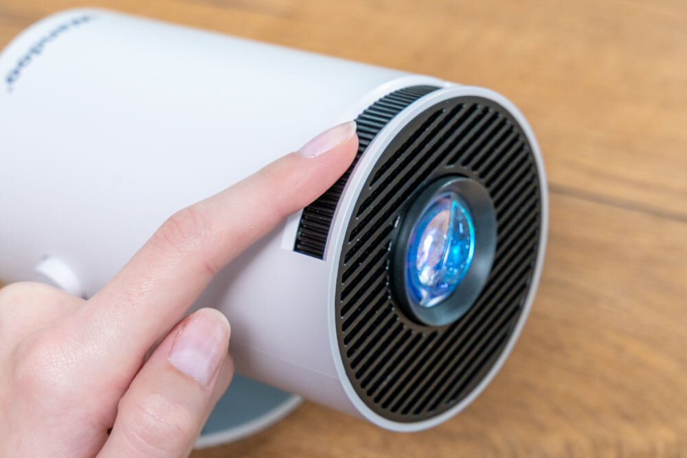 Projektor WANDOO LMP360 HD (1280 x 720), 160 ANSI lumen, Wi-Fi, Bluetooth Zbliżenie na przód projektora Wandoo. Palec kręci radełkowanym pierścieniem ostrości (regulacja ostrości) wokół soczewki projektora. Ręczna regulacja ostrości obrazu w projektorze Wandoo.