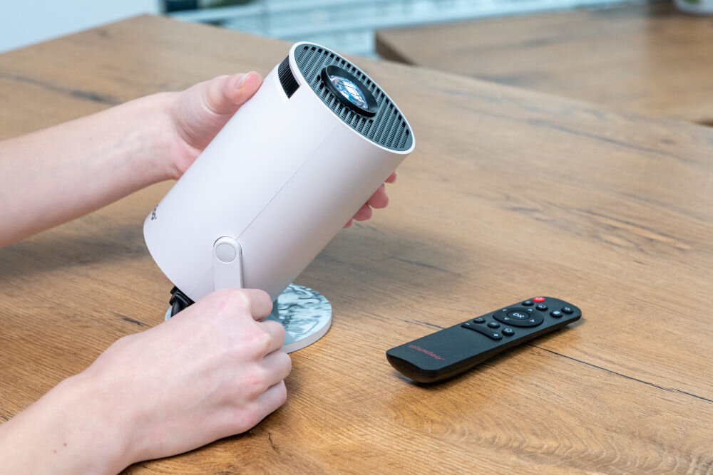 Projektor WANDOO LMP360 HD (1280 x 720), 160 ANSI lumen, Wi-Fi, Bluetooth Osoba trzyma oburącz kompaktowy, biały projektor Wandoo na drewnianym stole, regulując jego pozycję na okrągłej podstawce. Obok leży pilot. Regulacja ustawienia małego projektora Wandoo.