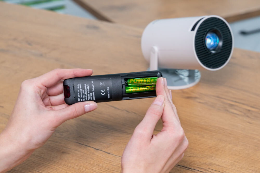 Projektor WANDOO LMP360 HD (1280 x 720), 160 ANSI lumen, Wi-Fi, Bluetooth Dłonie umieszczają dwie zielone baterie AAA 1,5V w komorze baterii czarnego pilota zdalnego sterowania projektora Wandoo. W tle projektor. Wymiana baterii AAA w pilocie projektora Wandoo.