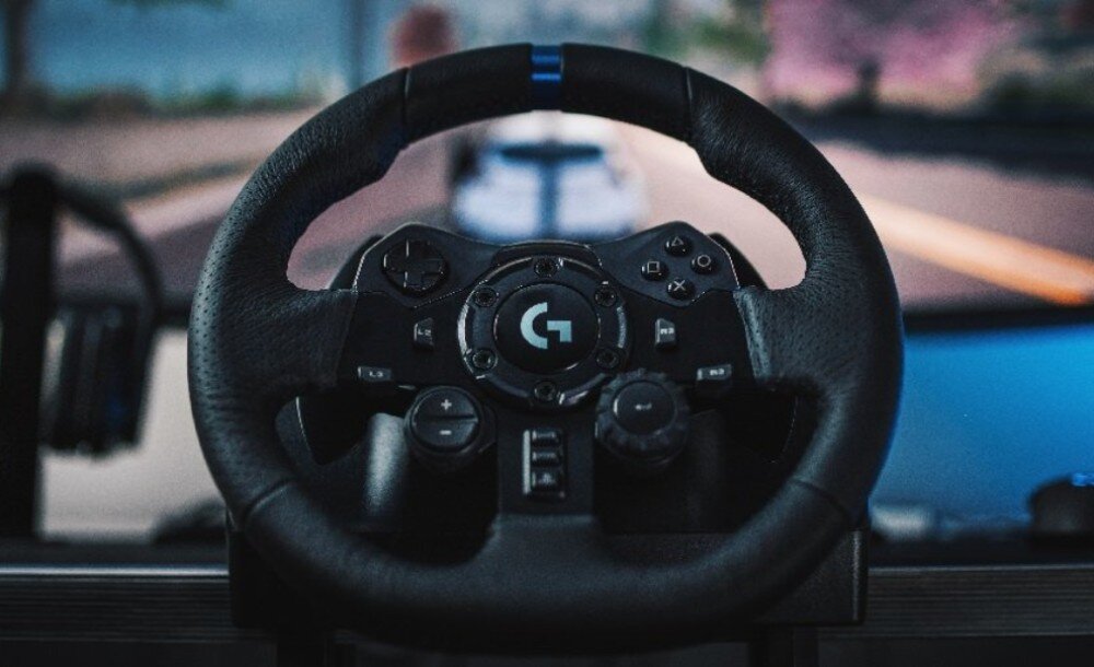 Kierownica LOGITECH G923 + Shifter Bundle Kierownica wyścigowa z przyciskami i pokrętłami zamontowana na biurku przed ekranem z widokiem na tor wyścigowy