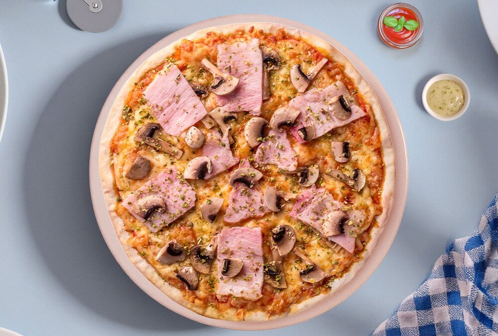 Na obrazie widoczna jest pizza z szynką i pieczarkami, umieszczona na kamieniu do pizzy. Obok znajdują się małe naczynia z sosem oraz serwetka w niebiesko-białą kratkę.