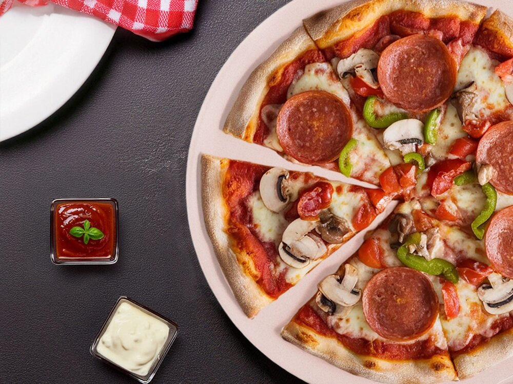 Na obrazie widoczna jest pizza z pepperoni, pieczarkami, papryką i serem, umieszczona na kamieniu do pizzy. Obok znajdują się dwa małe naczynia z sosem pomidorowym i białym sosem.