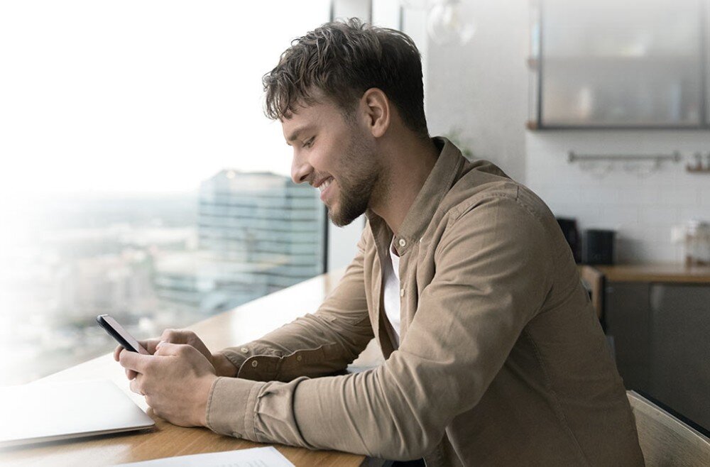 Router ASUS RT-BE58U Mężczyzna w beżowej koszuli uśmiechający się do ekranu telefonu siedząc przy drewnianym blacie