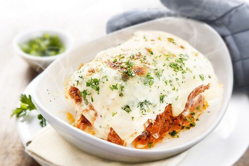 Na białym talerzu znajduje się porcja lasagne z widocznym serem i posypką z ziół. W tle widać szarą rękawicę kuchenną i małą miseczkę z zielonymi ziołami.