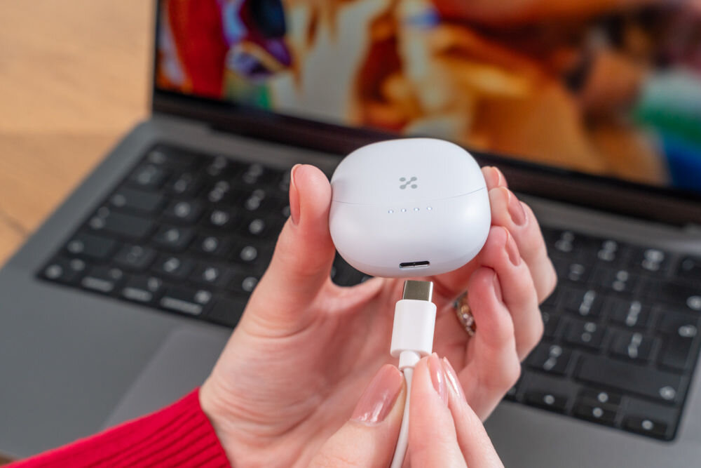 Słuchawki douszne XMUSIC XTW300B Niebieski Ujęcie z bliska, na którym dłonie podłączają biały kabel USB-C do portu ładowania w dolnej części białego, zaokrąglonego etui słuchawek. Etui ma na górze cztery diody LED, wskazujące stan naładowania. W tle klawiatura laptopa.