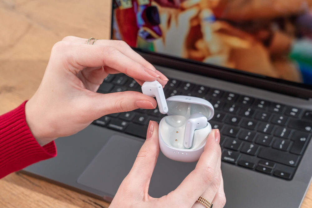 Słuchawki douszne XMUSIC XTW300B Niebieski Dłonie wyjmują jasnoniebieską słuchawkę douszną z otwartego, białego etui. Słuchawki znajdują się nad klawiaturą laptopa.