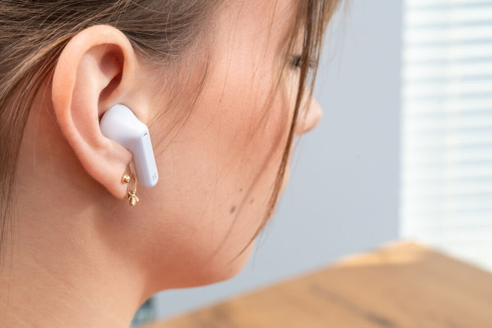 Słuchawki douszne XMUSIC XTW300B Niebieski Ujęcie z bliska na ucho kobiety, w którym znajduje się biała słuchawka douszna typu 'stem' (z pałąkiem). Słuchawka ma widoczne logo.