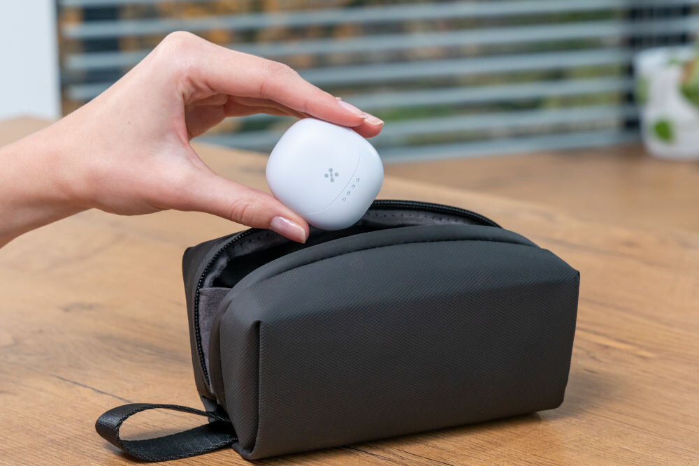 Słuchawki douszne XMUSIC XTW300B Niebieski Dłoń trzyma zamknięte, białe etui do słuchawek nad otwartym, czarnym etui/pokrowcem materiałowym. Na etui widoczne są cztery diody LED oraz logo.