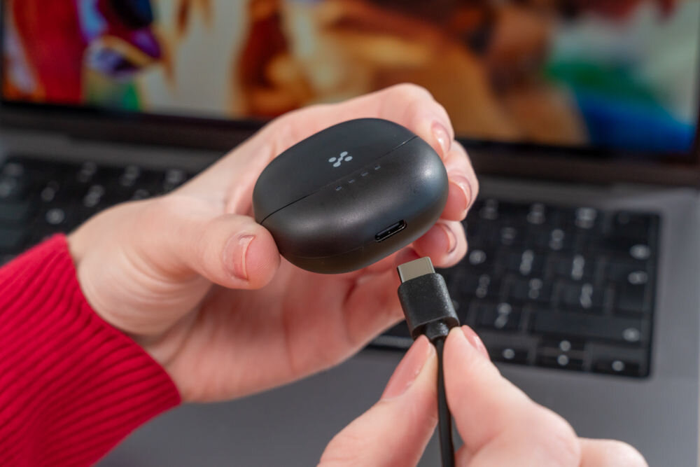 Słuchawki douszne XMUSIC XTW300B Czarny Ujęcie z bliska, na którym dłonie podłączają czarny kabel USB-C do portu ładowania w dolnej części czarnego etui słuchawek. Etui ma widoczne cztery białe diody LED stanu naładowania. W tle widać klawiaturę laptopa.