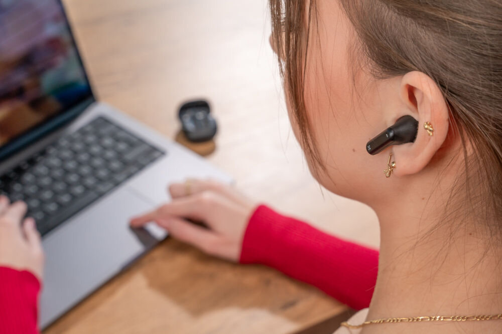 Słuchawki douszne XMUSIC XTW300B Czarny Ujęcie z bliska na ucho kobiety, w którym znajduje się czarna słuchawka douszna z pałąkiem (typu 'stem'). W tle kobieta pisze na laptopie.