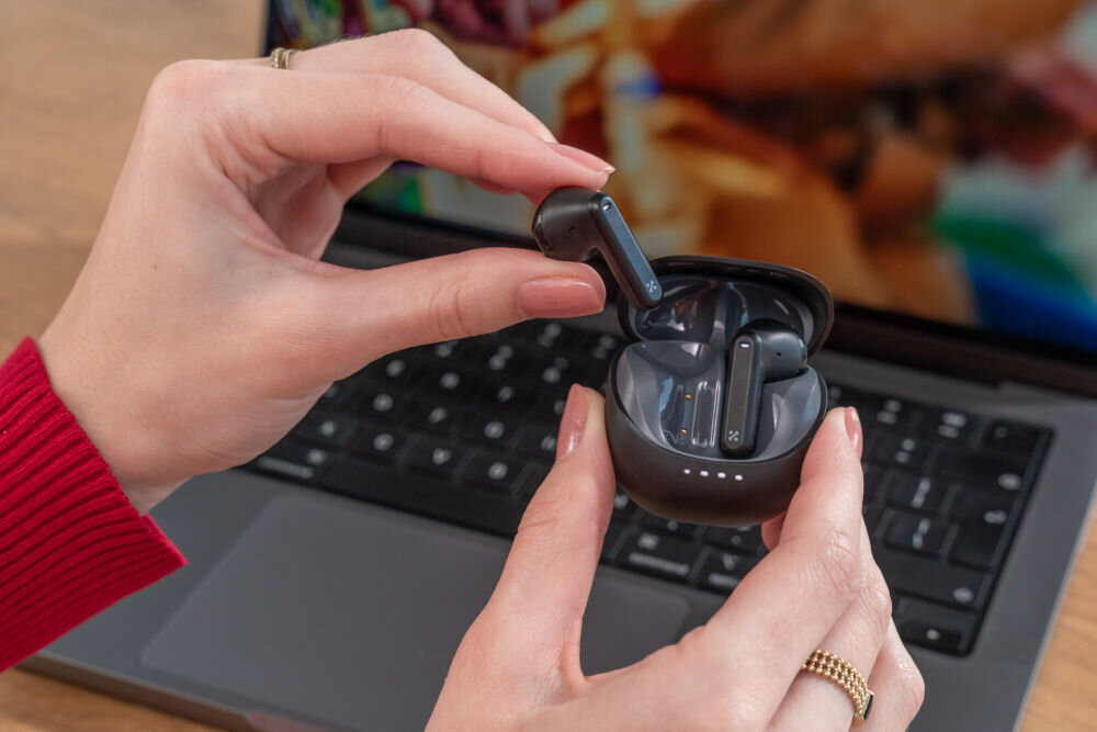 Słuchawki douszne XMUSIC XTW300B Czarny Dłonie wyjmują czarną słuchawkę douszną z otwartego, czarnego etui. Słuchawki znajdują się nad klawiaturą laptopa.