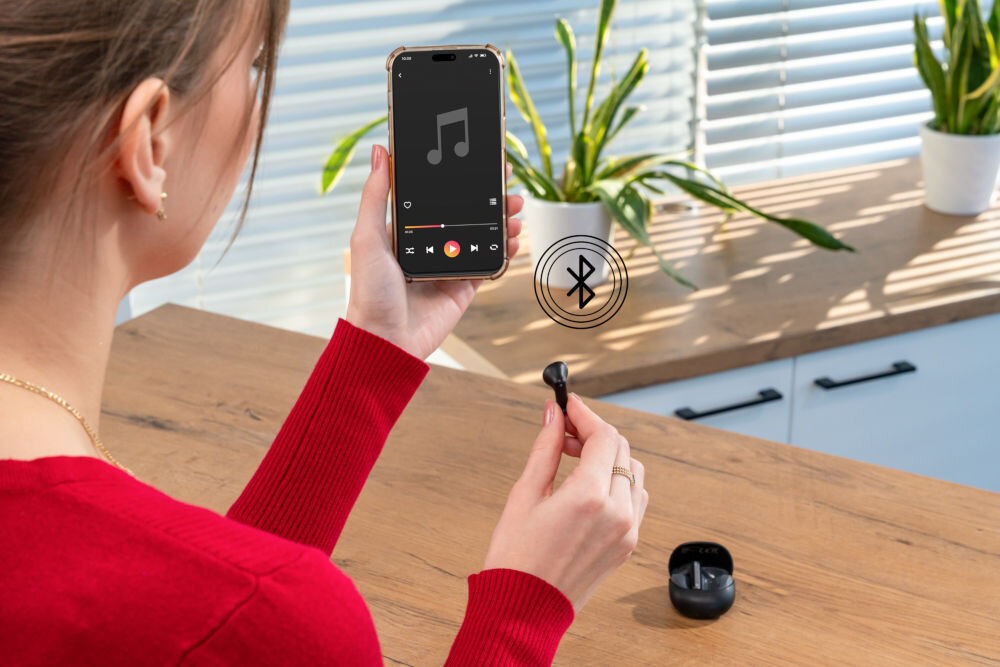 Słuchawki douszne XMUSIC XTW300B Czarny Kobieta trzyma w prawej ręce smartfon z ekranem odtwarzacza muzyki, a w lewej ręce jedną czarną słuchawkę. Ikonka Bluetooth symbolizuje bezprzewodowe połączenie. Otwarty futerał jest na blacie.