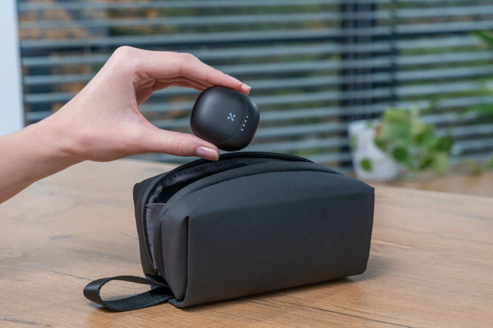 Słuchawki douszne XMUSIC XTW300B Czarny Dłoń trzyma zamknięte, czarne etui do słuchawek z widocznym logo i diodami, unosząc je nad otwartym, czarnym pokrowcem materiałowym (saszetką).