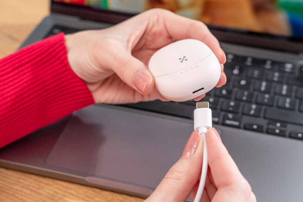 Słuchawki douszne XMUSIC XTW300P różowy Ujęcie z bliska, na którym dłonie podłączają różowy kabel USB-C do portu ładowania w dolnej części różowego etui słuchawek. Etui ma widoczne cztery białe diody LED stanu naładowania. W tle widać klawiaturę laptopa.
