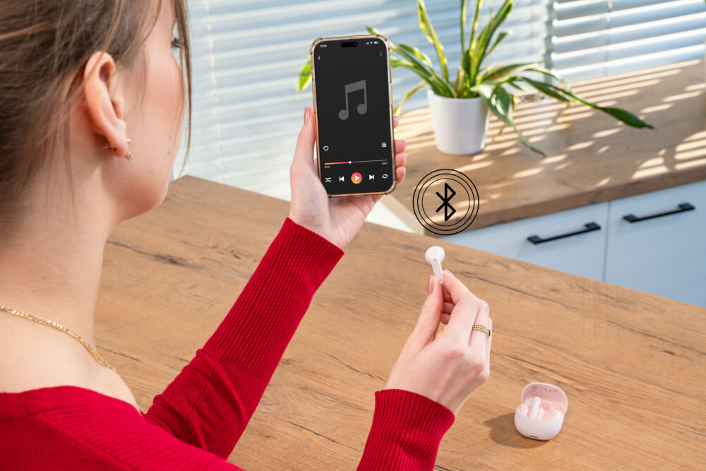 Słuchawki douszne XMUSIC XTW300P różowy Kobieta trzyma w prawej ręce smartfon z ekranem odtwarzacza muzyki, a w lewej ręce jedną różową słuchawkę. Ikonka Bluetooth symbolizuje bezprzewodowe połączenie. Otwarty futerał jest na blacie.