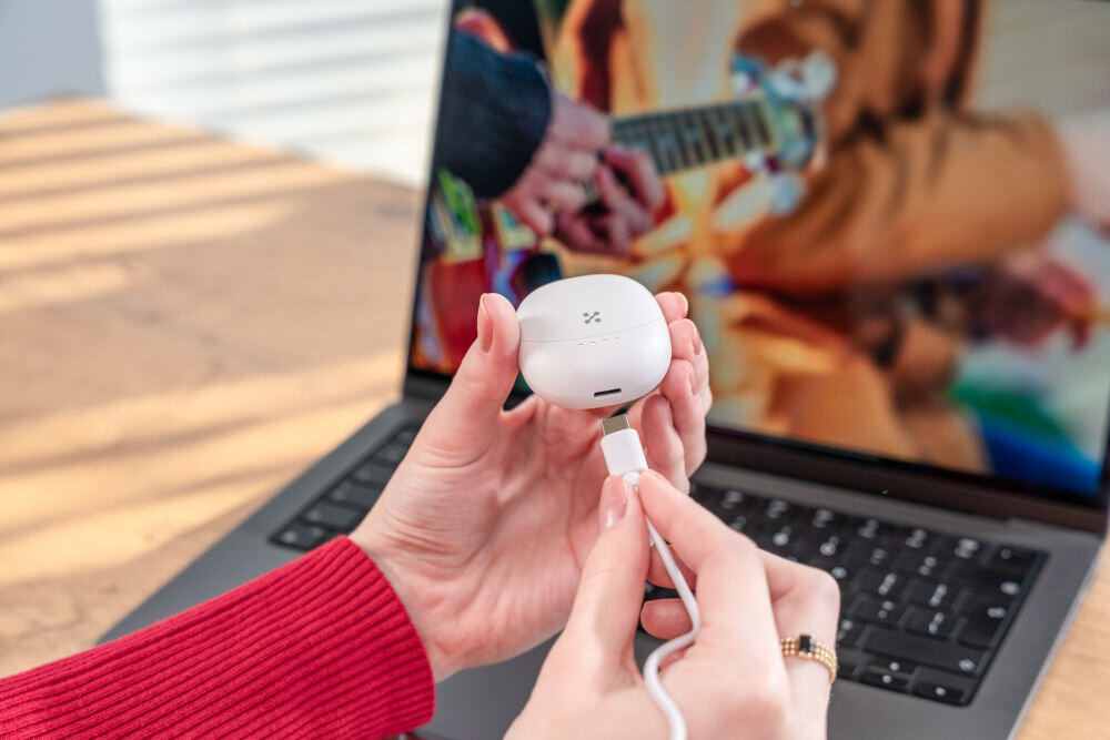 Słuchawki douszne XMUSIC XTW300W biały Ujęcie z bliska, na którym dłonie podłączają biały kabel USB-C do portu ładowania w dolnej części białego etui słuchawek. Etui ma widoczne cztery białe diody LED stanu naładowania. W tle widać klawiaturę laptopa.