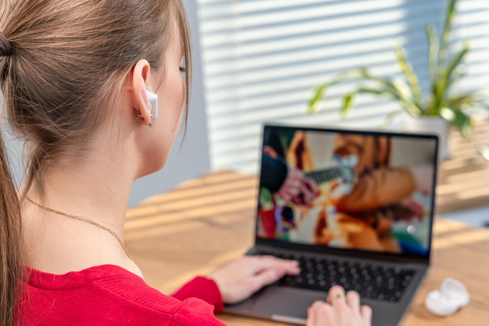 Słuchawki douszne XMUSIC XTW300W biały Ujęcie z bliska na ucho kobiety, w którym znajduje się biała słuchawka douszna z pałąkiem (typu 'stem'). W tle kobieta pisze na laptopie.