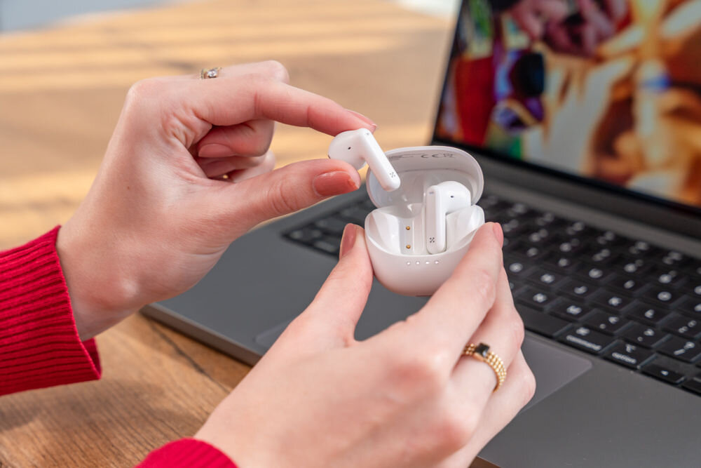 Słuchawki douszne XMUSIC XTW300W biały Dłonie wyjmują białą słuchawkę douszną z otwartego, czarnego etui. Słuchawki znajdują się nad klawiaturą laptopa.