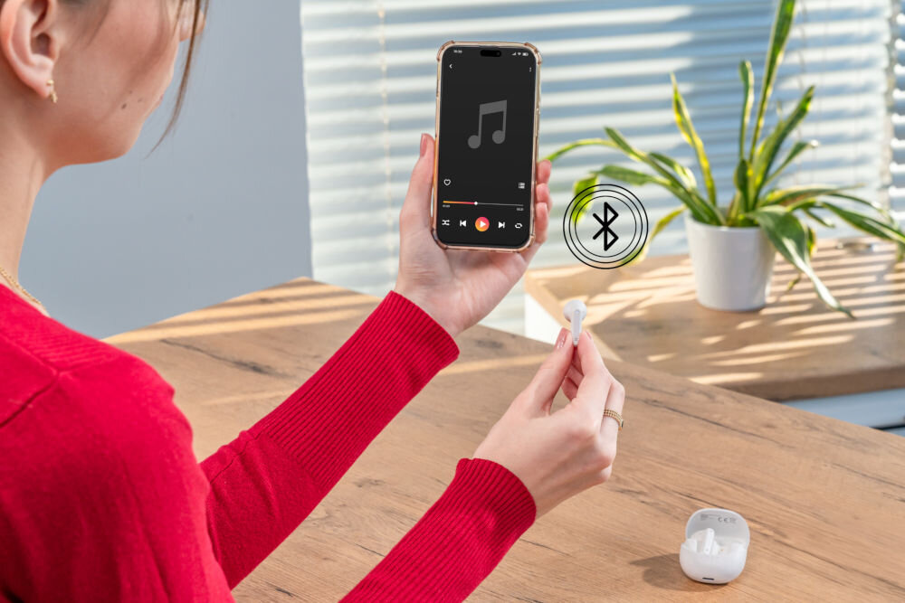 Słuchawki douszne XMUSIC XTW300W biały Kobieta trzyma w prawej ręce smartfon z ekranem odtwarzacza muzyki, a w lewej ręce jedną białą słuchawkę. Ikonka Bluetooth symbolizuje bezprzewodowe połączenie. Otwarty futerał jest na blacie.