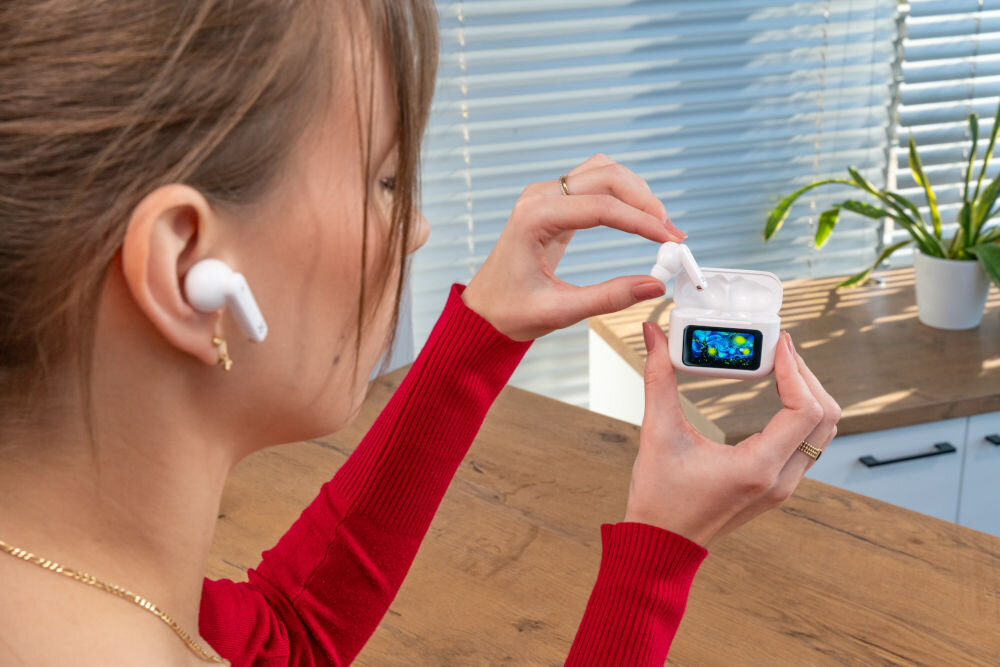 Słuchawki dokanałowe XMUSIC XTW950W ANC Biały Młoda kobieta w czerwonym swetrze, z włożoną jedną słuchawką do ucha, wyjmuje drugą słuchawkę z otwartego etui ze Smart Screen. Scena dzieje się w kuchni/biurze z widokiem na żaluzje i blat.