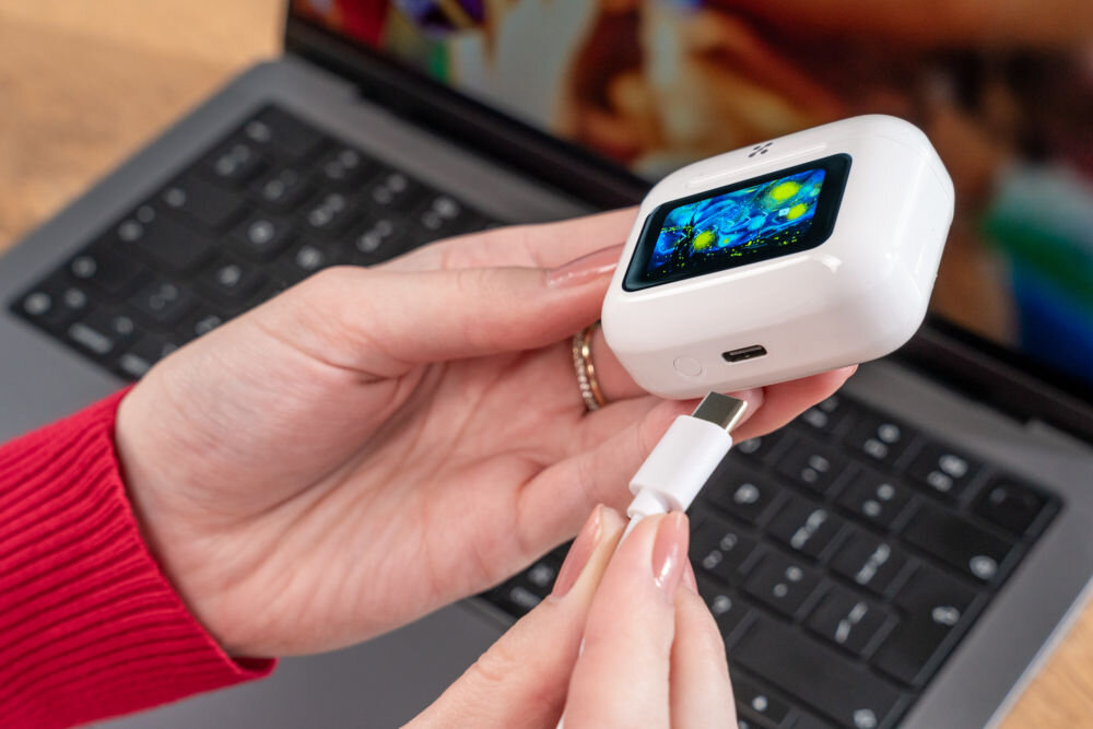 Słuchawki dokanałowe XMUSIC XTW950W ANC Biały Ujęcie z bliska, na którym dłonie podłączają biały kabel USB-C do portu ładowania w dolnej części białego etui słuchawek ze Smart Screen. W tle widać klawiaturę laptopa.