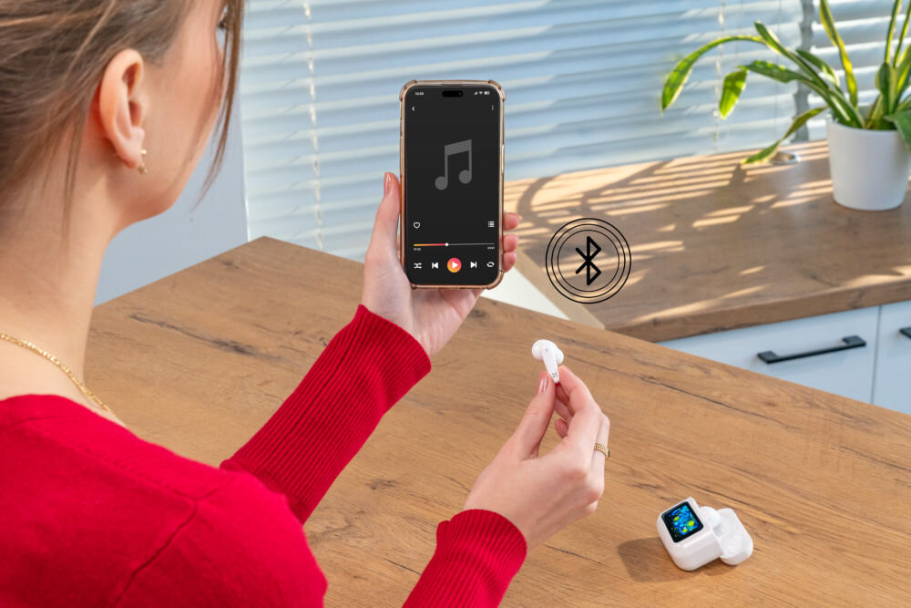 Słuchawki dokanałowe XMUSIC XTW950W ANC Biały Kobieta trzyma w lewej ręce smartfon z ekranem odtwarzacza muzyki, a w prawej ręce jedną słuchawkę. Obok symbolizuje to ikonka Bluetooth, wskazując na bezprzewodowe połączenie. Etui ze Smart Screen leży na drewnianym blacie.