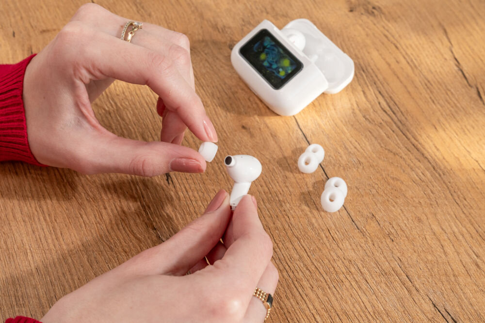 Słuchawki dokanałowe XMUSIC XTW950W ANC Biały Ujęcie z bliska na dłonie wymieniające silikonową nakładkę (gumkę) na białej słuchawce dousznej. Obok leżą dwie dodatkowe nakładki i etui ze Smart Screen.