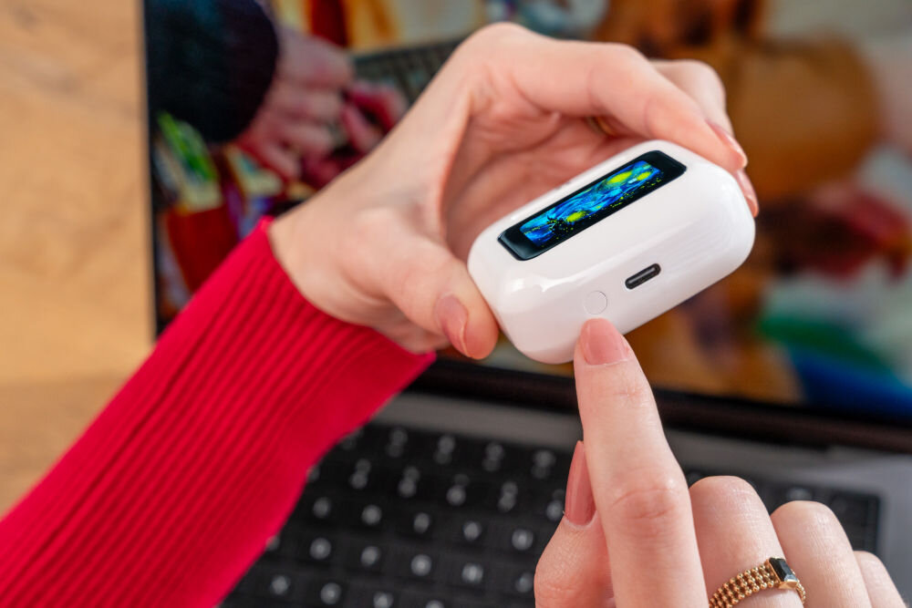 Słuchawki dokanałowe XMUSIC XTW950W ANC Biały Ujęcie z bliska na spód białego etui do słuchawek ze Smart Screen. Palec wskazuje na mały okrągły przycisk funkcyjny, obok którego znajduje się port USB-C.