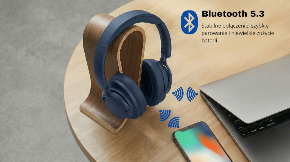 Na drewnianym stojaku znajdują się niebieskie słuchawki nauszne. Obok widoczny jest telefon, laptop oraz tekst: \'Bluetooth 5.3 Stabilne połączenie, szybkie parowanie i niewielkie zużycie baterii\'.