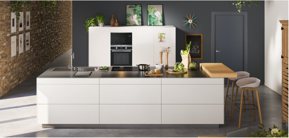 Piekarnik FRANKE Smart FSM 86 H BK Zdjęcie przedstawia kuchnię z dużą wyspą, na której znajdują się warzywa, deska do krojenia oraz naczynia. W zabudowie ściennej widoczny jest zestaw piekarnika i kuchenki mikrofalowej, tworzący spójną, elegancką aranżację