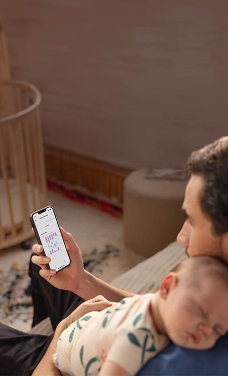 Mężczyzna ze śpiącym niemowlęciem, trzymający w dłoni telefon z aplikacją Baby Monitor+