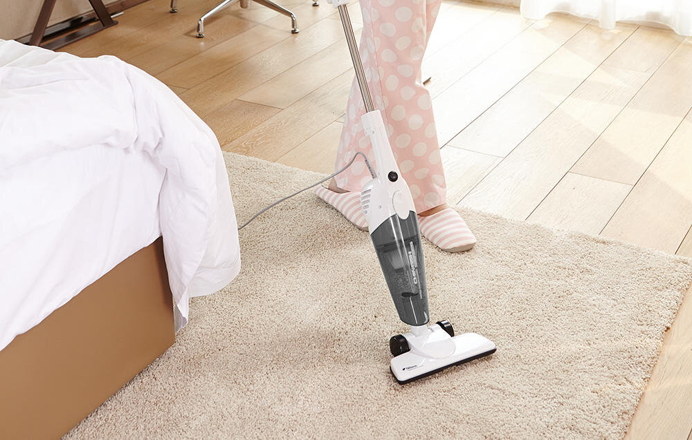 Na obrazie widoczny jest biało-szary odkurzacz pionowy na dywanie, obok łóżka. W tle widać osobę w piżamie w kropki oraz drewnianą podłogę.