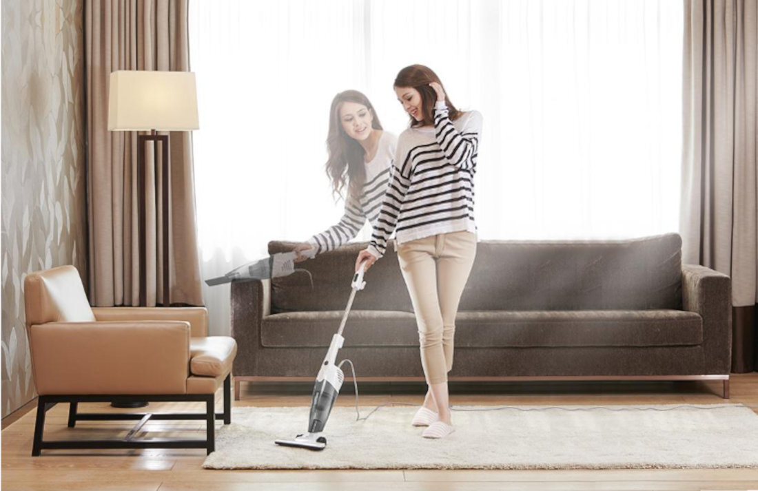 Osoba używa odkurzacza pionowego w jasnym salonie z sofą, lampą i fotelem. Odkurzacz ma biało-szary kolor i jest używany na dywanie.