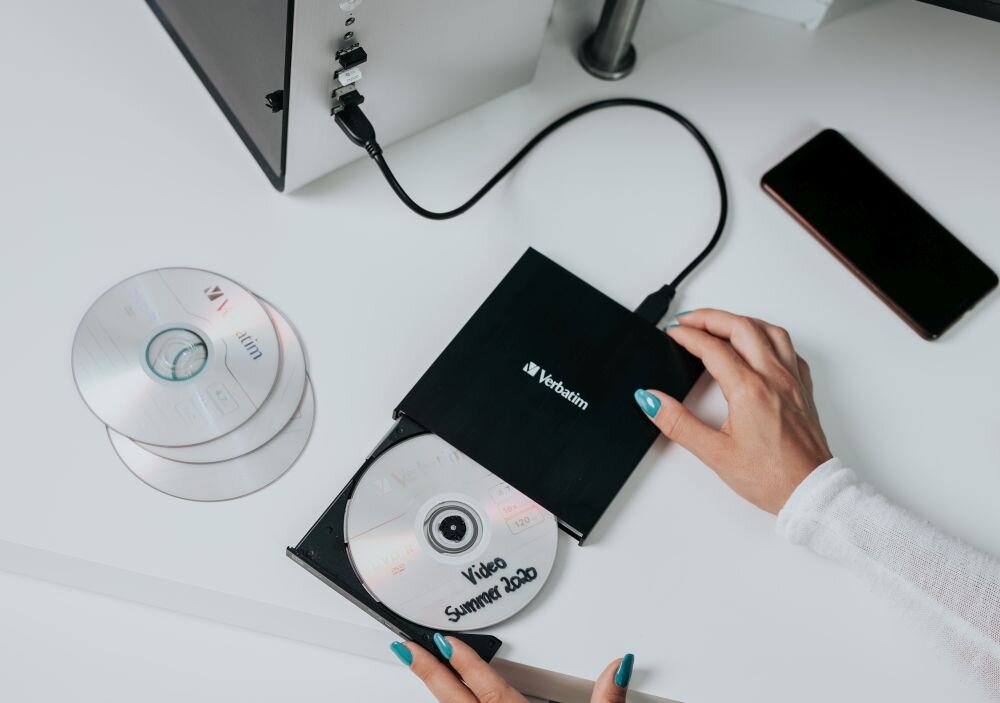 Nagrywarka VERBATIM External Slimline CD/DVD Writer Czarny napęd optyczny podłączony kablem, w wysuniętej tacce znajduje się płyta z napisem 'Video Summer 2020', obok leżą dwie płyty i smartfon przenośna nagrywarka USB