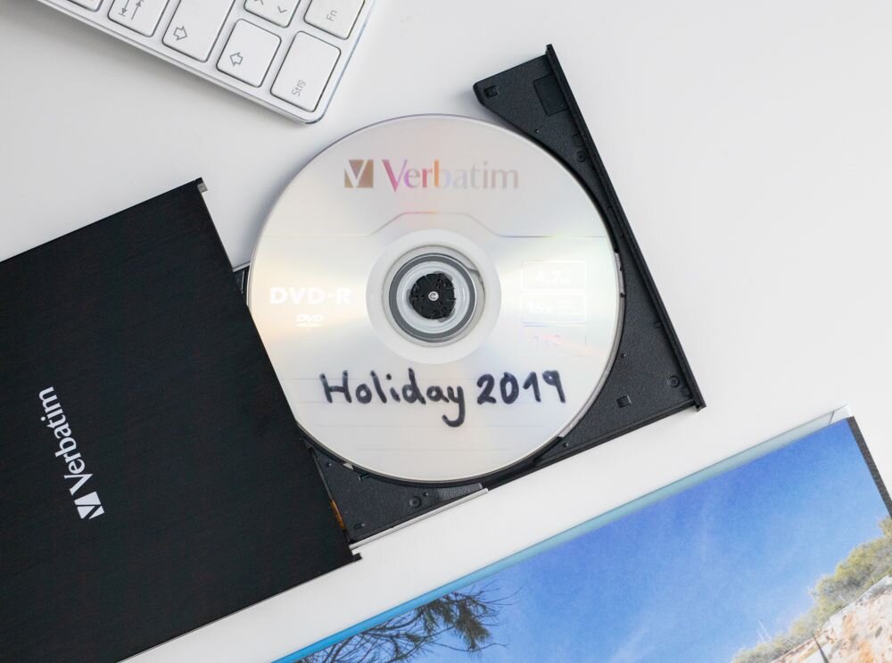 Nagrywarka VERBATIM External Slimline CD/DVD Writer Zbliżenie na płytę w wysuniętej tacce czarnego napędu, na płycie widoczny napis 'Holiday 2019', obok fragment klawiatury i album ze zdjęciem plaży zasilanie z USB, idealna do laptopów