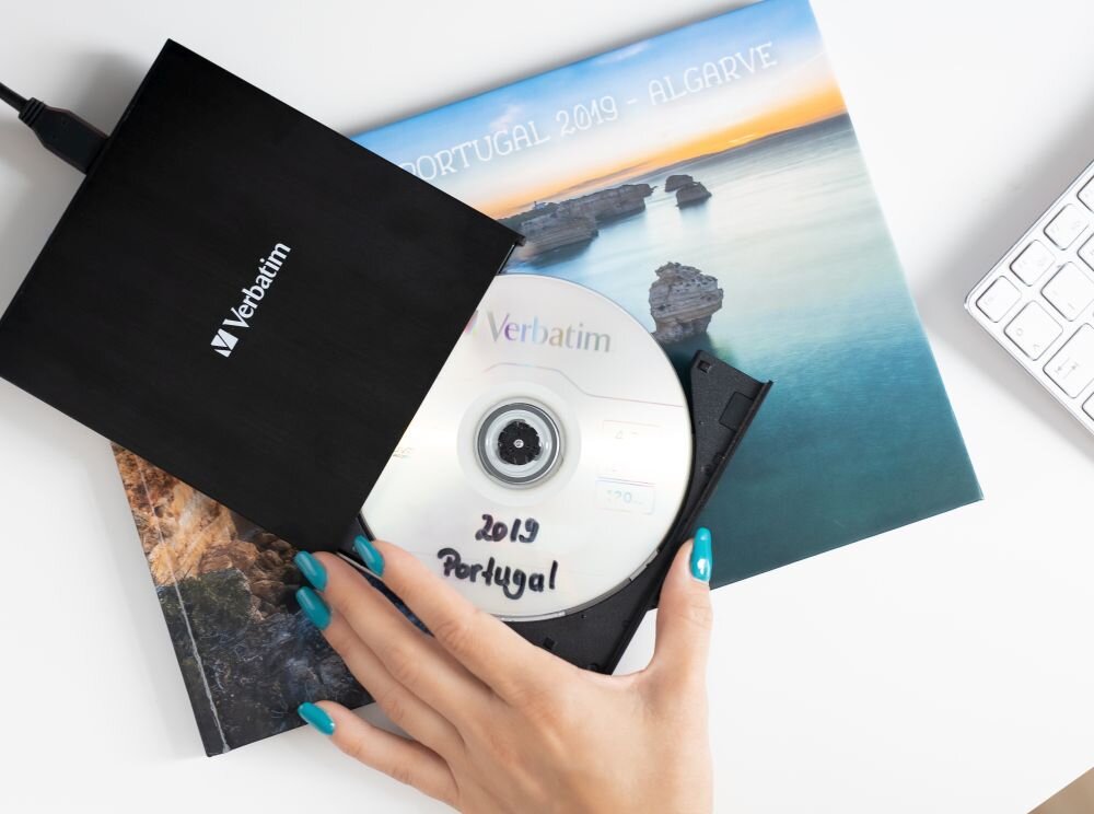Nagrywarka VERBATIM External Slimline CD/DVD Writer Czarny napęd optyczny z wysuniętą tacką, w środku znajduje się płyta z napisem '2019 Portugal', pod napędem leży album z okładką przedstawiającą krajobraz nadmorski, obok fragment klawiatury nagrywarka CD/DVD kompatybilna z Windows i macOS