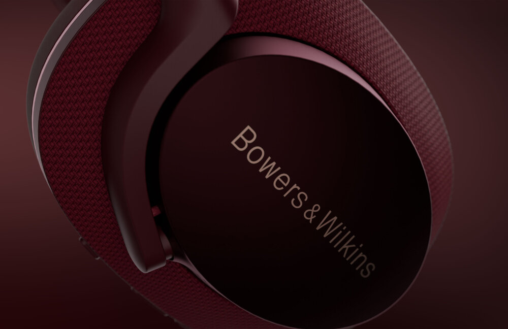 Osoba nosi czerwone słuchawki nauszne z widocznym napisem \'Bowers & Wilkins\'. W tle rozmyte drzewa i budynki.