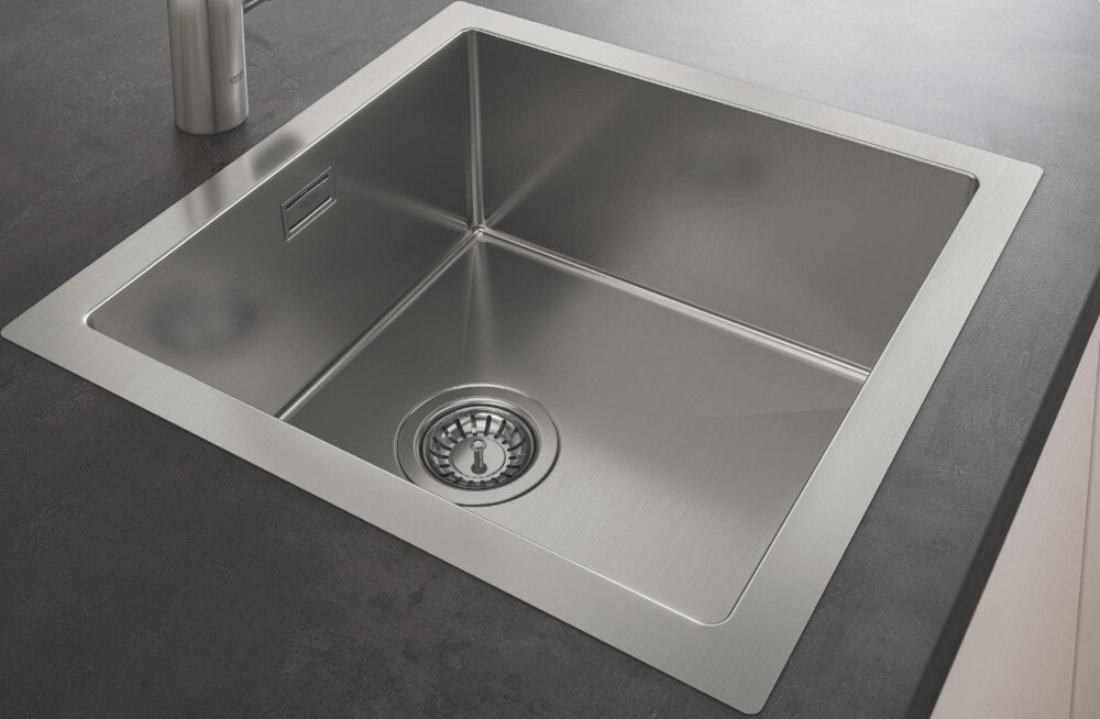 Zlewozmywak jednokomorowy GROHE K700 31578SD1 Stal szlachetna Na zdjęciu widać stalowy zlewozmywak jednokomorowy z ociekaczem, osadzony w blacie o ciemnoszarej powierzchni. W centralnej części ociekacza znajduje się chromowana, jednouchwytowa bateria stojąca, a w tle widoczne są minimalistyczne akcesoria kuchenne na białej półce.