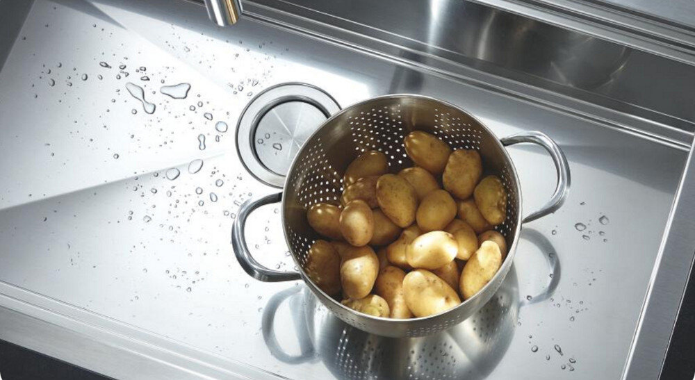 Zlewozmywak jednokomorowy GROHE K700 31578SD1 Na zdjęciu na stalowym ociekaczu zlewozmywaka stoi metalowy durszlak wypełniony gorącymi ziemniakami w łupinach. Wokół sitka widoczne są krople wody oraz fragment wylewki baterii, co podkreśla odporność powierzchni na wysoką temperaturę Stal szlachetna odporność na wysokie temperatury stal nierdzewna gorące naczynia odporność termiczna trwały zlew kuchenny
