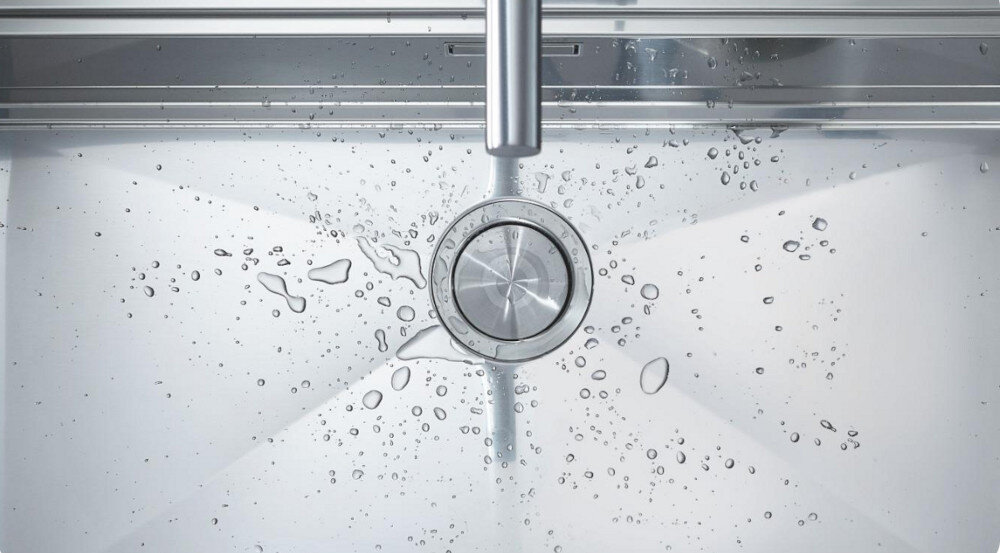 Zlewozmywak jednokomorowy GROHE K700 31578SD1 Stal szlachetna Na zdjęciu z góry pokazana jest stalowa komora zlewozmywaka z centralnie umieszczonym, okrągłym odpływem i promieniście opadającym dnem. Wokół odpływu widoczne są rozpryski i krople wody oraz fragment wylewki baterii, co podkreśla precyzyjne kształtowanie powierzchni cichy zlewozmywak redukcja hałasu GROHE Whisper tłumienie dźwięku komfort użytkowania