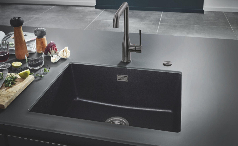 Zlewozmywak jednokomorowy GROHE K700U 31655AP0 Czarny jednokomorowy zlewozmywak wpuszczany w blat z wysoką, chromowaną baterią kuchenną. Wokół stoją produkty spożywcze i dodatki kuchenne tworzące estetyczną aranżację