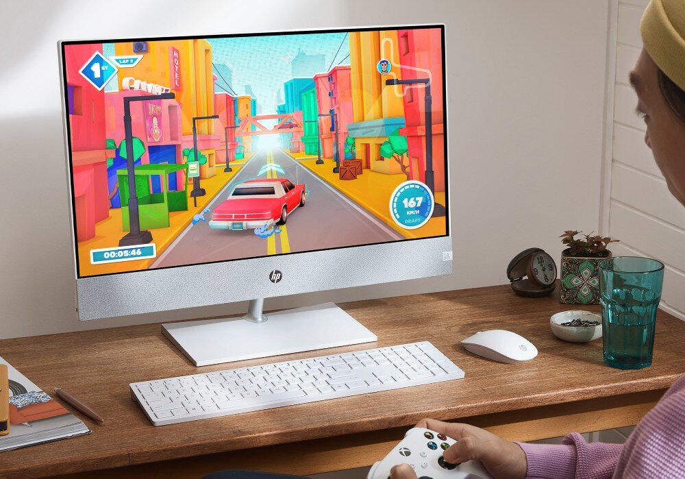 Komputer HP Pavilion 27-ca2092nw Osoba siedzi przy biurku i gra w kolorową grę wyścigową na komputerze typu all-in-one z białą klawiaturą i myszą na drewnianym blacie, opis