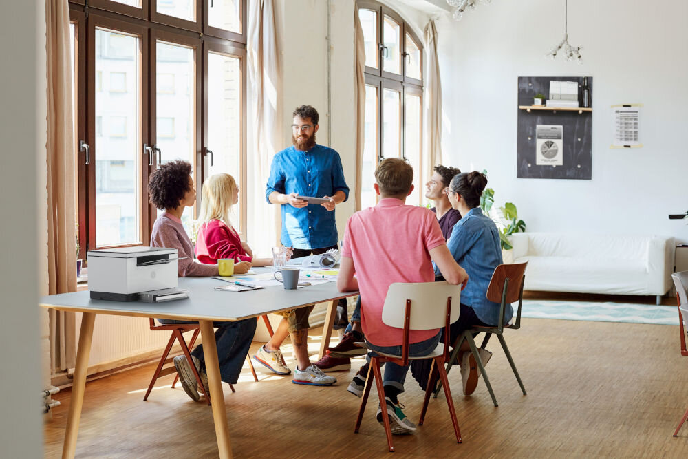 Urządzenie wielofunkcyjne HP LaserJet M234d Automatyczny druk dwustronny czarno-białe wydruki, prezentacja urządzenia na stole ludzie przy nim siedzący