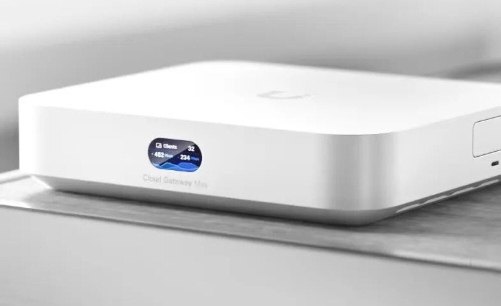 Router UBIQUITI UCG-MAX-EU Białe urządzenie sieciowe o zaokrąglonych rogach leżące na szarej powierzchni, z małym wyświetlaczem na froncie pokazującym liczbę klientów i prędkość transmisji